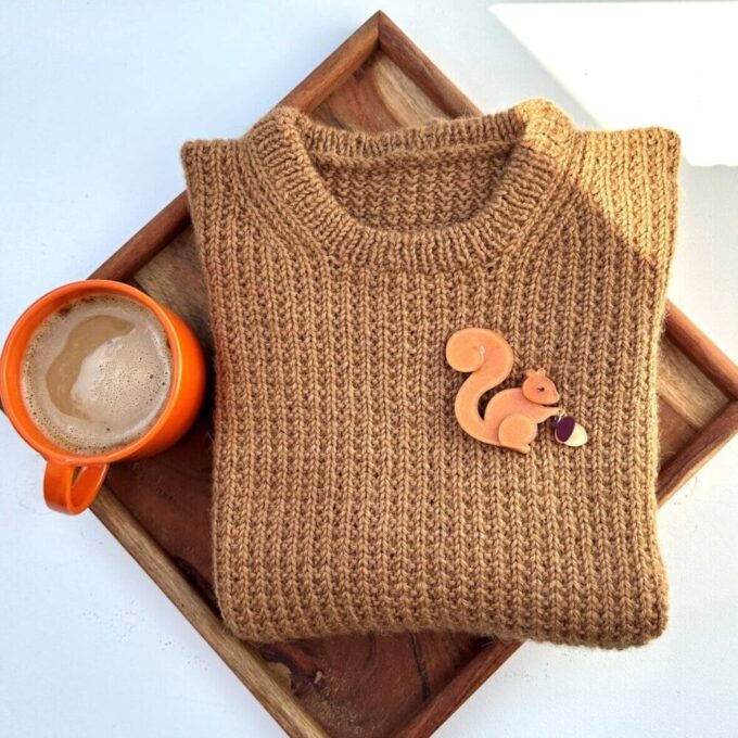 Bakke med orange keramikkop med kaffe, en nystrikket Friday Slipover og en broche med et egern